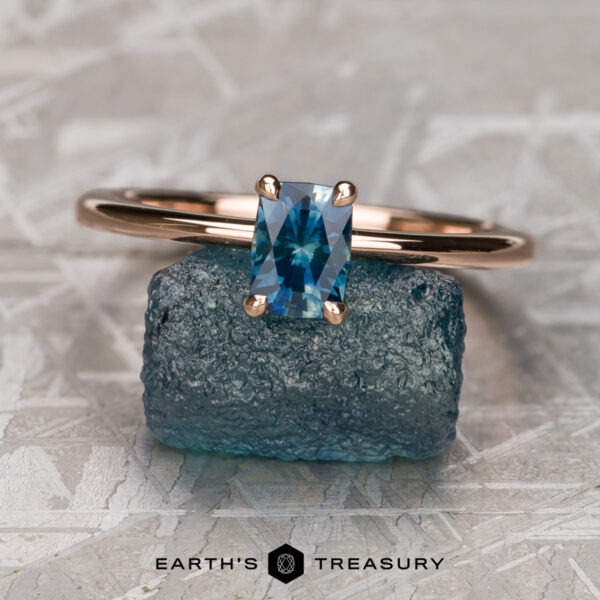 The "Flora" ring in 14K rose gold with 0.68-carat Montana sapphire The "Flora" ring in 14K rose gold with a 0.68-carat Montana sapphire is displayed on a raw blue stone, reminiscent of treasures from Flora Solitaire, with "Earth's Treasury" written below.