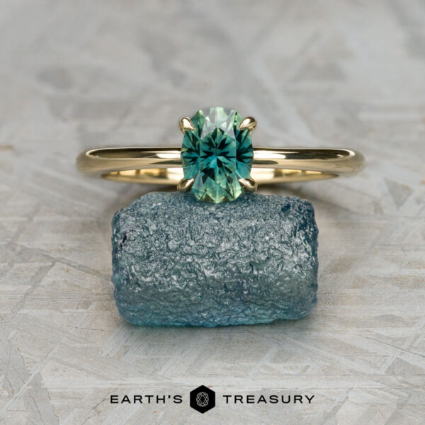 The "Flora" ring in 14k yellow gold with 1.04-carat Australian sapphire The "Flora" ring in 14k yellow gold with a 1.04-carat Australian sapphire stands upright on rough blue stone, echoing Flora Solitaire’s elegance, with "EARTH'S TREASURY" text and logo at the bottom center.