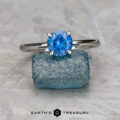 The "Flora" ring in platinum with a 1.20-carat Montana sapphire is displayed on a rough blue stone, with the "EARTH'S TREASURY" text and logo at the bottom.