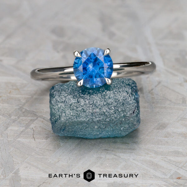 The "Flora" ring in platinum with 1.20-carat Montana sapphire The "Flora" ring in platinum with a 1.20-carat Montana sapphire is displayed on a rough blue stone, with the "EARTH'S TREASURY" text and logo at the bottom.