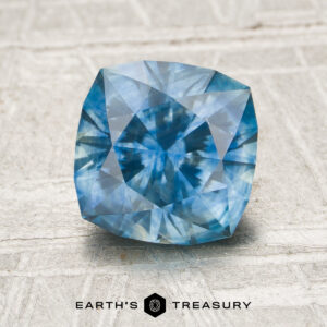 A faceted Montana Sapphire, product MS1762, with visible internal patterns is displayed on a light textured surface. The Earth's Treasury logo appears below the gem.