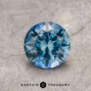 A 1.32-Carat Robin’s Egg Blue Montana Sapphire (Heated) is displayed on a textured gray surface with "EARTH’S TREASURY" at the bottom.
