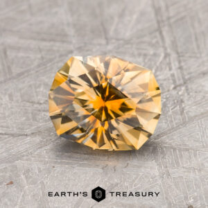 A 1.59-carat Yellow-Orange Particolored Montana Sapphire (Heated) is shown on a textured gray surface with the “Earth’s Treasury” logo and text visible below.