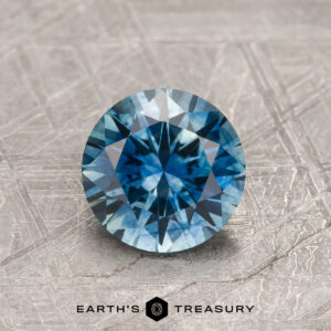 A 1.17-carat teal blue Montana Sapphire (heated), round brilliant-cut, stands upright on a textured gray surface with the "Earth's Treasury" logo beneath it.