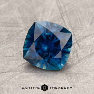 A 1.54-carat deep peacock Montana sapphire (heated), square-cut, rests on a textured gray surface with "EARTH'S TREASURY" and a geometric logo below.