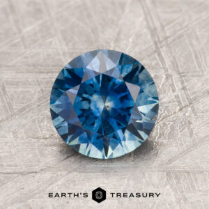 A 1.61-carat Peacock Blue Montana Sapphire (Heated), round brilliant-cut, sits on a textured gray surface, with the "EARTH'S TREASURY" logo and text at the bottom.