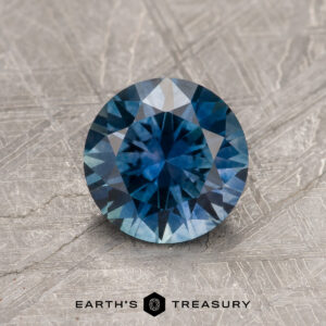 A 1.24-Carat Peacock Blue Montana Sapphire (Heated) shines in vivid blue on a textured metallic background, with the text "Earth's Treasury" beneath.