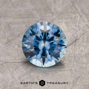 The 1.27-Carat Rich Sky Blue Montana Sapphire (Heated) is showcased in a brilliant round cut on a textured gray background, with the "Earth's Treasury" logo and text displayed below.
