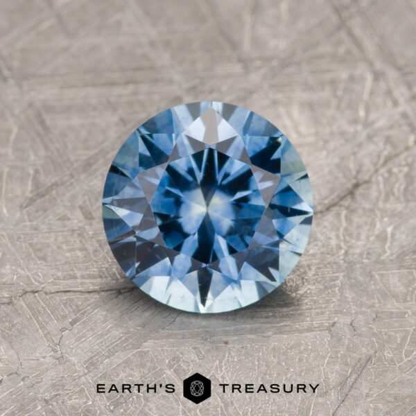 1.27-Carat Rich Sky Blue Montana Sapphire (Heated) The 1.27-Carat Rich Sky Blue Montana Sapphire (Heated) is showcased in a brilliant round cut on a textured gray background, with the "Earth's Treasury" logo and text displayed below.