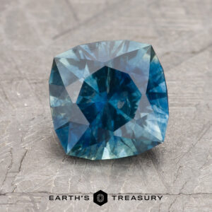 A 2.28-carat deep blue-green Montana sapphire (heated), cushion-cut, displayed on a textured gray surface with "EARTH'S TREASURY" text at the bottom.