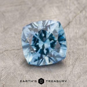 A 1.63-carat heated aqua blue Montana Sapphire with a cushion cut sits on a textured gray surface, showing light and dark blue facets. The text "Earth's Treasury" appears at the bottom.
