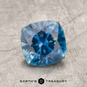 A 1.61-Carat Medium Blue Montana Sapphire (Heated), cushion cut and faceted, rests on a textured gray surface with the "EARTH'S TREASURY" logo below.