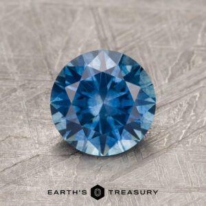 1.27-carat rich teal blue Montana sapphire (heated), round brilliant-cut, displayed on a textured gray surface with "Earth's Treasury" logo and text at the bottom.