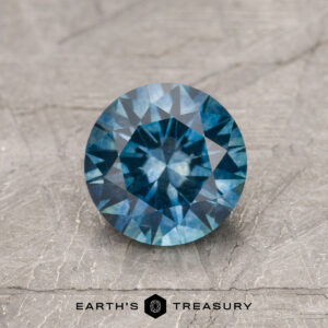 A 1.30-Carat Teal Blue Montana Sapphire (Heated), round brilliant-cut, sits on a textured grey surface displaying "EARTH'S TREASURY" and a logo beneath.