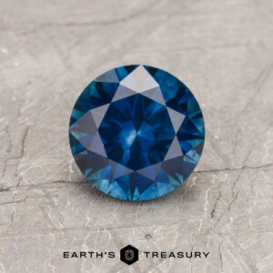 A 1.14-carat deep peacock blue Montana sapphire (heated), round brilliant-cut, is displayed on a textured gray surface with "EARTH'S TREASURY" at the bottom.
