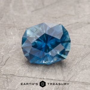 A 1.58-carat oval-cut Peacock Blue Montana Sapphire (heated) with multiple facets is displayed on a textured gray surface, featuring the text "EARTH'S TREASURY" at the bottom.