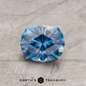 A 1.39-carat medium blue heated Montana Sapphire with geometric facets is displayed on a textured gray surface, with "Earth's Treasury" branding below.