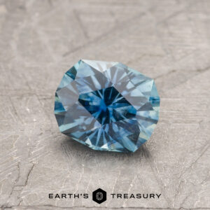 A 1.33-carat Teal Blue Montana Sapphire (Heated), round and faceted, is shown on a textured gray surface with "EARTH'S TREASURY" written at the bottom.