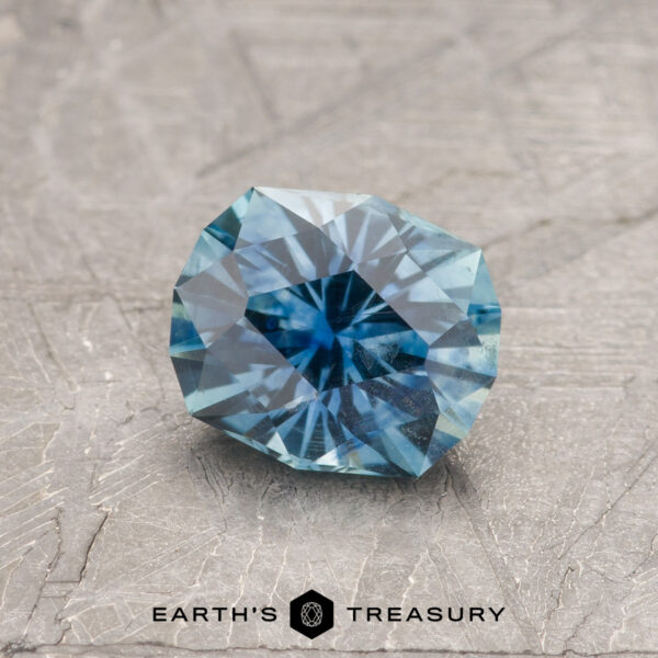1.33-Carat Teal Blue Montana Sapphire (Heated) A 1.33-carat Teal Blue Montana Sapphire (Heated), round and faceted, is shown on a textured gray surface with "EARTH'S TREASURY" written at the bottom.