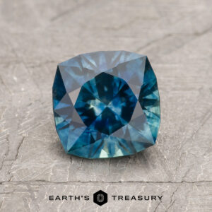 A 1.49-carat deep blue-green Montana Sapphire (heated) with a cushion cut is displayed on a textured gray surface, featuring the text "Earth's Treasury" beneath it.