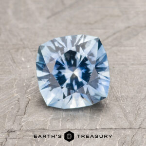 A 1.43-carat sky blue Montana sapphire (heated), cushion-cut, is showcased on a textured gray surface with "Earth's Treasury" displayed below.