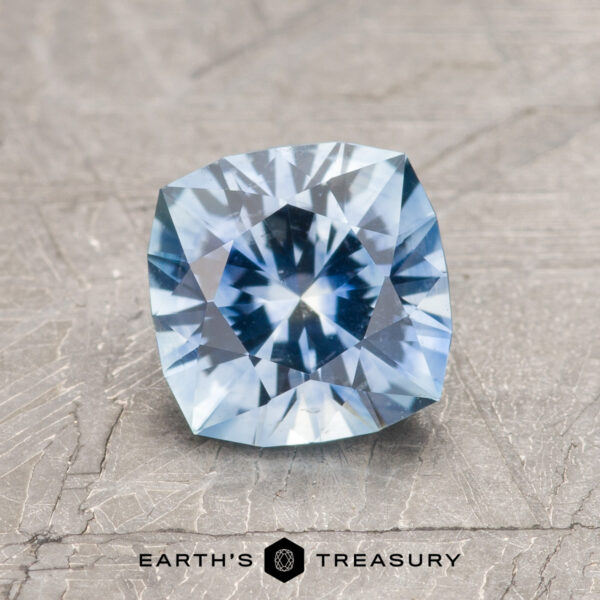 1.43-Carat Sky Blue Montana Sapphire (Heated) A 1.43-carat sky blue Montana sapphire (heated), cushion-cut, is showcased on a textured gray surface with "Earth's Treasury" displayed below.