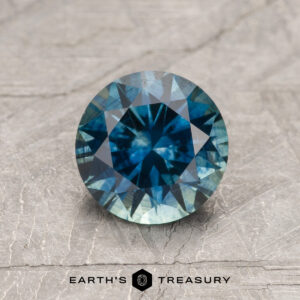 A 1.29-carat Rich Teal Montana Sapphire (Heated) gemstone, round-cut, is shown on a textured gray surface with "EARTH'S TREASURY" displayed beneath it.