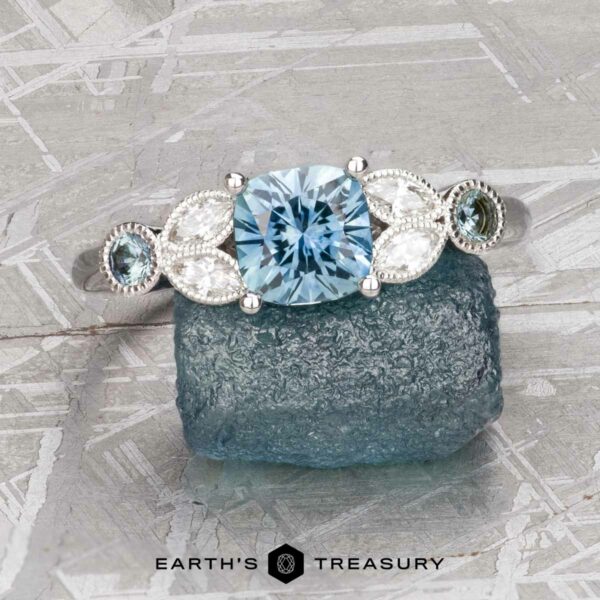 The "Calypso" ring in 14K white gold with 1.20-carat Montana sapphire The "Calypso" ring in 14K white gold with a 1.20-carat cushion-cut Montana sapphire and side stones is shown on a rough blue stone against a geometric backdrop. "Earth's Treasury" appears below.