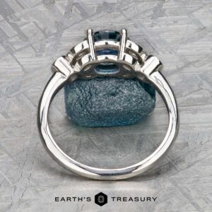 The "Calypso" ring in platinum with a 2.10-carat Montana sapphire stands upright before a rough blue stone on textured gray, with the "Earth's Treasury" logo at the bottom.