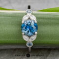 The "Calypso" ring in platinum showcases a 2.10-carat Montana sapphire, accented by smaller blue stones and marquise-shaped white details, elegantly displayed on a green plant leaf.