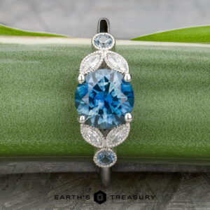 The "Calypso" ring in platinum showcases a 2.10-carat Montana sapphire, accented by smaller blue stones and marquise-shaped white details, elegantly displayed on a green plant leaf.