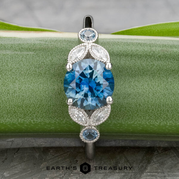 The "Calypso" ring in platinum with 2.10-carat Montana sapphire The "Calypso" ring in platinum showcases a 2.10-carat Montana sapphire, accented by smaller blue stones and marquise-shaped white details, elegantly displayed on a green plant leaf.