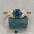 The "Calypso" ring in 18K yellow gold features a 1.47-carat Montana sapphire center, accented by marquise and round diamonds, displayed on a raw blue stone with the “Earth’s Treasury” logo below.