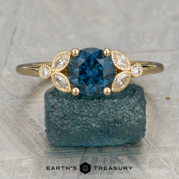 The "Calypso" ring in 18K yellow gold with 1.47-carat Montana sapphire The "Calypso" ring in 18K yellow gold features a 1.47-carat Montana sapphire center, accented by marquise and round diamonds, displayed on a raw blue stone with the “Earth’s Treasury” logo below.