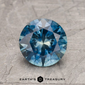 A 2.20-carat Peacock Blue-Yellow Particolored Heated Montana Sapphire is shown on a textured gray surface with the "Earth's Treasury" logo and text below it.