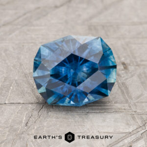 A 1.91-carat rich blue Montana Sapphire (heated), cut in a faceted oval shape, rests on a textured gray surface with the "Earth's Treasury" logo below.