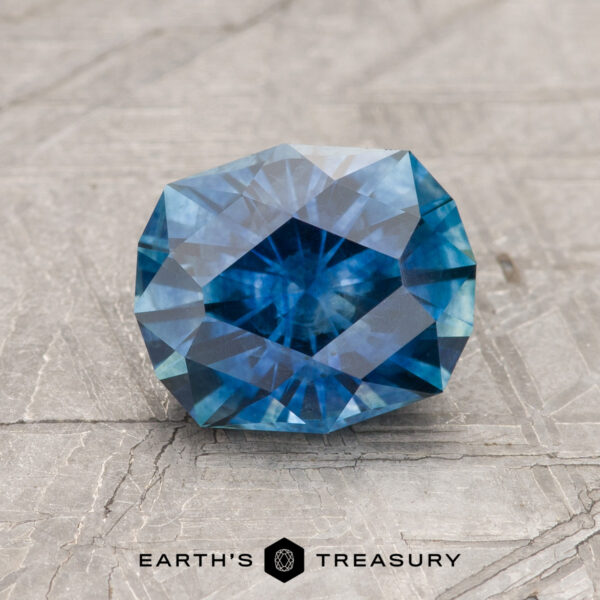1.91-Carat Rich Blue Montana Sapphire (Heated) A 1.91-carat rich blue Montana Sapphire (heated), cut in a faceted oval shape, rests on a textured gray surface with the "Earth's Treasury" logo below.