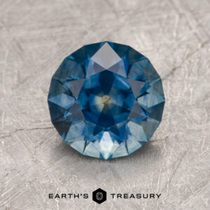 A 1.97-carat deep blue Montana sapphire (heated) in a round brilliant cut is displayed on a textured gray surface, with the "Earth's Treasury" logo and text below.