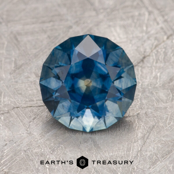 1.97-Carat Deep Blue Montana Sapphire (Heated) A 1.97-carat deep blue Montana sapphire (heated) in a round brilliant cut is displayed on a textured gray surface, with the "Earth's Treasury" logo and text below.