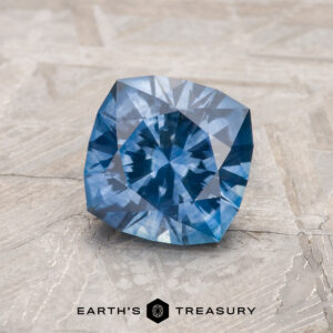 The faceted Medium Blue Montana Sapphire, product MS2237, rests on a textured light surface with "Earth's Treasury" and a hexagon logo shown below.