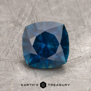 A 2.23-carat Deep Peacock Blue Montana Sapphire (Heated) in cushion cut is shown on a textured gray surface with "EARTH'S TREASURY" text and logo beneath.