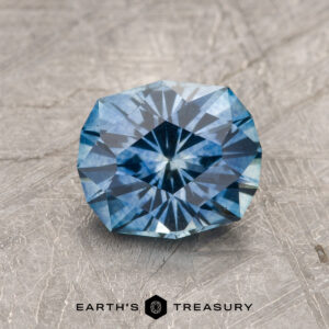 A 1.82-carat medium blue heated Montana Sapphire is showcased on a textured gray surface with "EARTH'S TREASURY" displayed at the bottom.