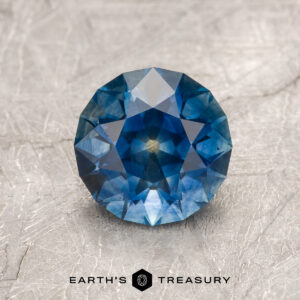 A 1.79-carat rich blue Montana Sapphire (heated) in round brilliant cut rests on a textured, light surface with the "Earth's Treasury" logo below.