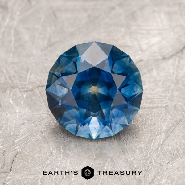 1.79-Carat Rich Blue Montana Sapphire (Heated) A 1.79-carat rich blue Montana Sapphire (heated) in round brilliant cut rests on a textured, light surface with the "Earth's Treasury" logo below.