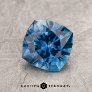 A 1.95-carat medium blue Montana Sapphire (heated), cushion-cut with geometric facets, sits on a textured gray surface. "Earth's Treasury" appears at the bottom of the image.