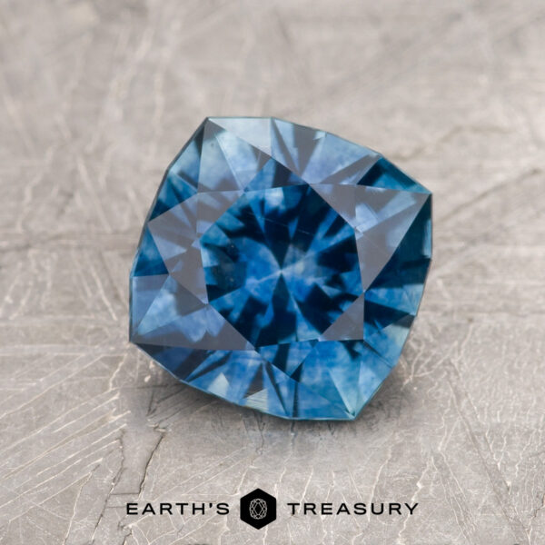 1.95-Carat Medium Blue Montana Sapphire (Heated) A 1.95-carat medium blue Montana Sapphire (heated), cushion-cut with geometric facets, sits on a textured gray surface. "Earth's Treasury" appears at the bottom of the image.