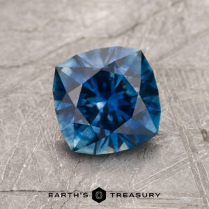 A 1.43-carat rich blue heated Montana Sapphire with intricate facets rests on a textured gray surface; "Earth's Treasury" appears below the gemstone.