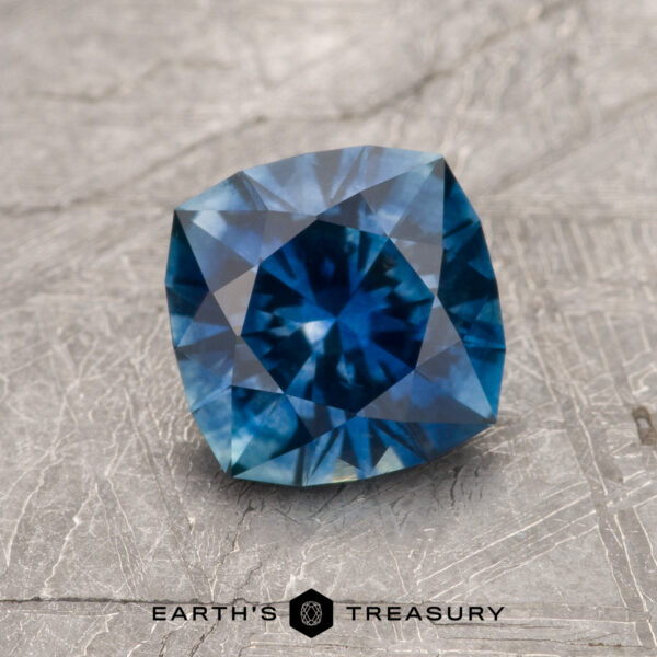 1.43-Carat Rich Blue Montana Sapphire (Heated) A 1.43-carat rich blue heated Montana Sapphire with intricate facets rests on a textured gray surface; "Earth's Treasury" appears below the gemstone.