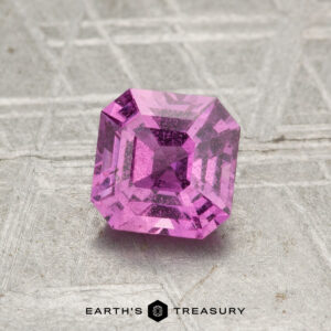 A 1.06-Carat Hot Pink Sapphire (Heated) with visible inclusions rests on a textured gray surface, with “EARTH’S TREASURY” displayed at the bottom.