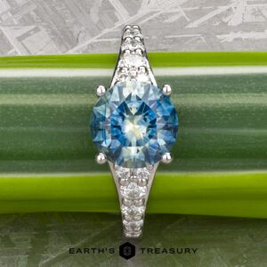 The "Arethusa" ring in platinum features a 2.65-carat round Montana sapphire and smaller clear stones along the band, shown on a green leaf with "Earth's Treasury" below—an exquisite, nature-inspired jewelry design.